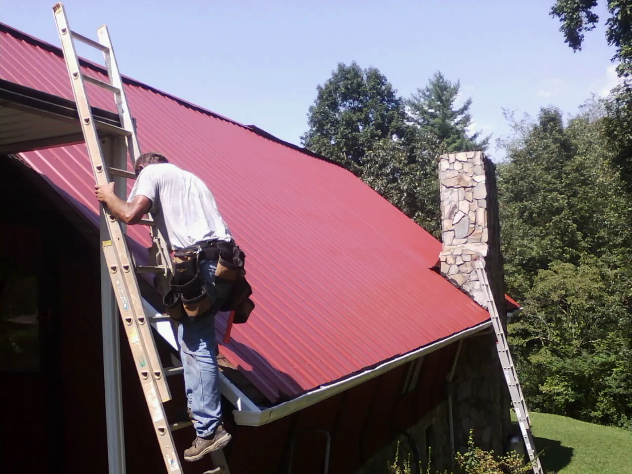 Roofer on a ladder beside a red metal roof and stone chimney in Immaculata